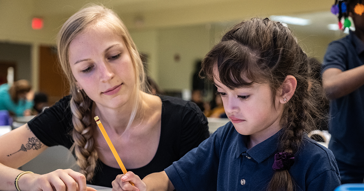 An adult looks over a students work while the student writes on paper with a pencil.