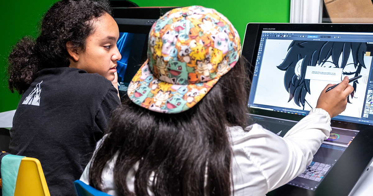 Two students working at a computer and pointing at a character drawing on the screen.