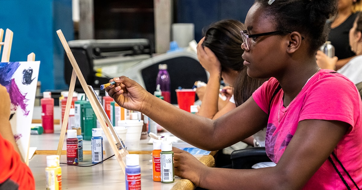 Students holds a paintbrush in front of a tabletop easily, while surrounded by paint bottles