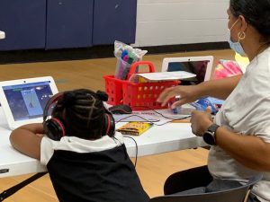 A girl wearing headphones looks at her virtual schoolwork in a gym, while an adult sits next to her.