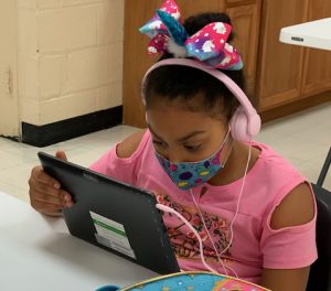 A girl wearing a hair bow, headphones and a masks looks at a screen while doing virtual schoolwork.